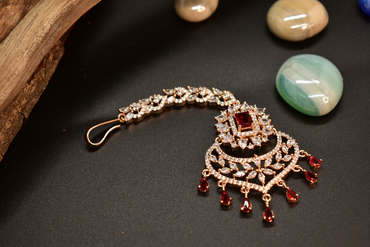 AD rosegold mang tikka with red gemstone in the middle and Ad drops displayed on a dark background