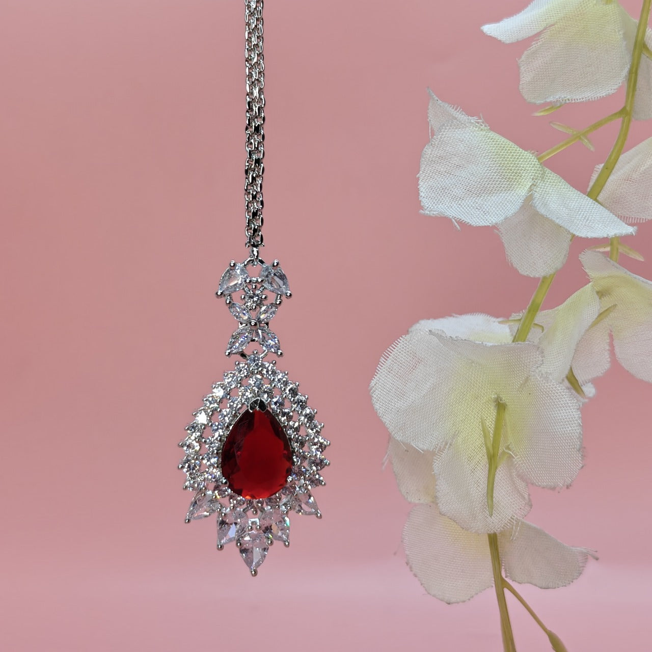American Diamond Elegant Mangtikka with a red stone and pink background with white flowers.