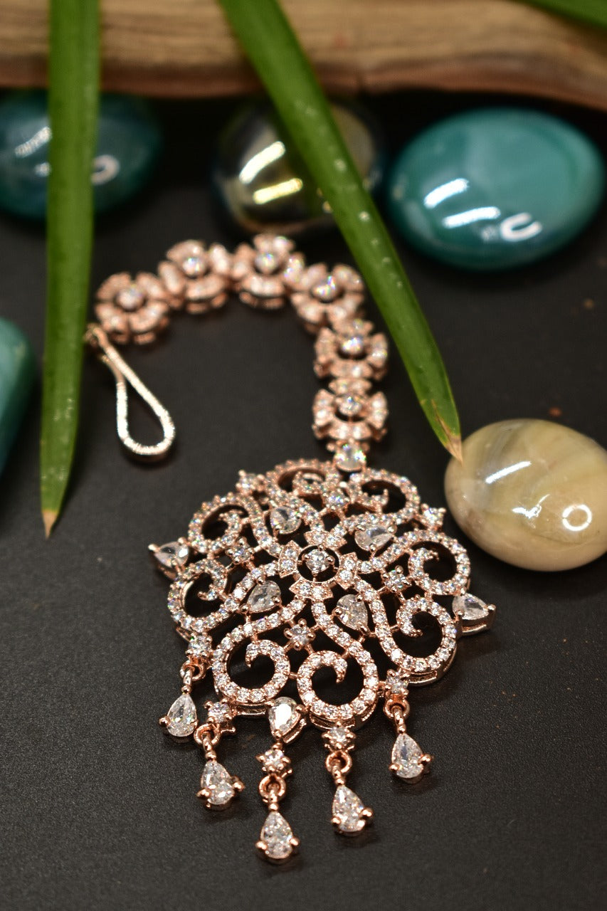Ornate rose gold Mangtikka with gemstones on a dark surface with green leaves and stones.
