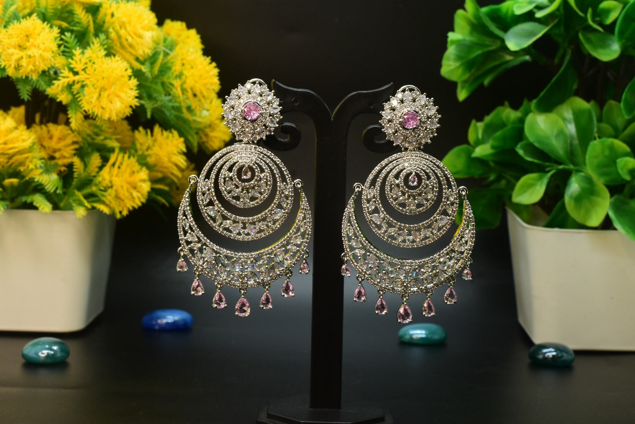 AD Pair of ornate chandelier earrings with pink gemstones on a stand with flowers and plants in the background