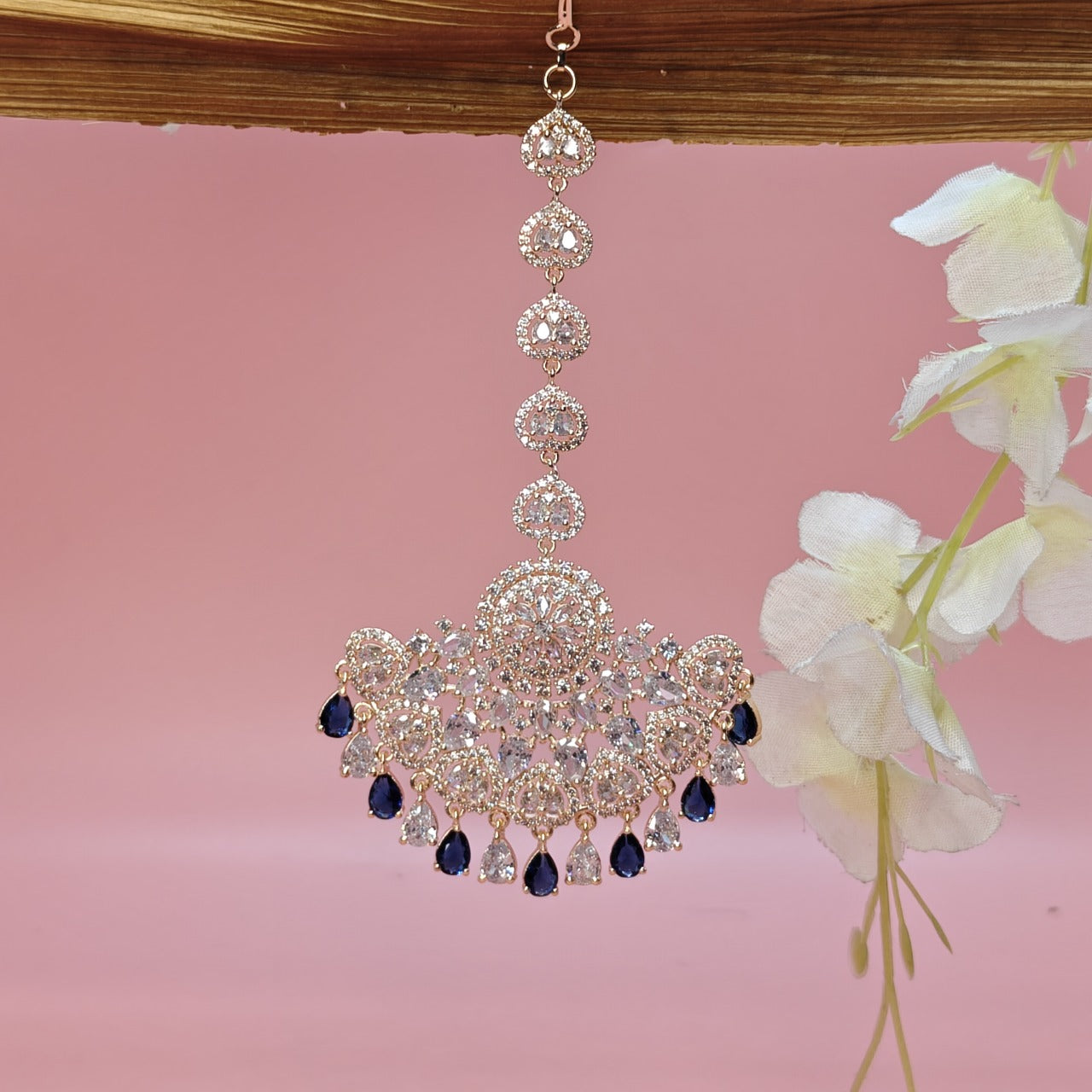 Elegant AD chandelier-style Mangtikka with blue accents on a pink background with flowers.