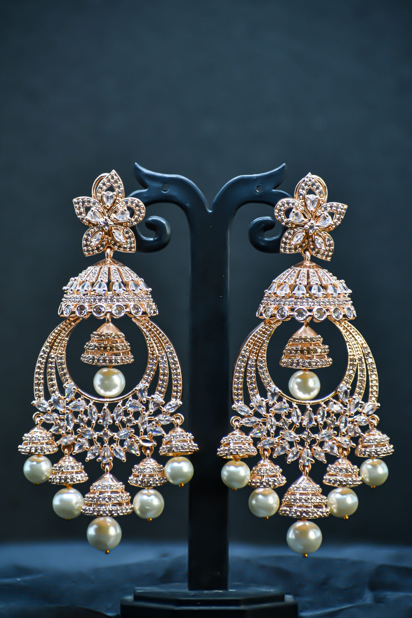 A pair of rose gold-plated zirconia jhumka earrings displayed on a stand against a black background.