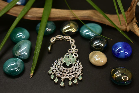 AD Silver plated Mang tikka with green gemstones on a dark surface with stones and leaves