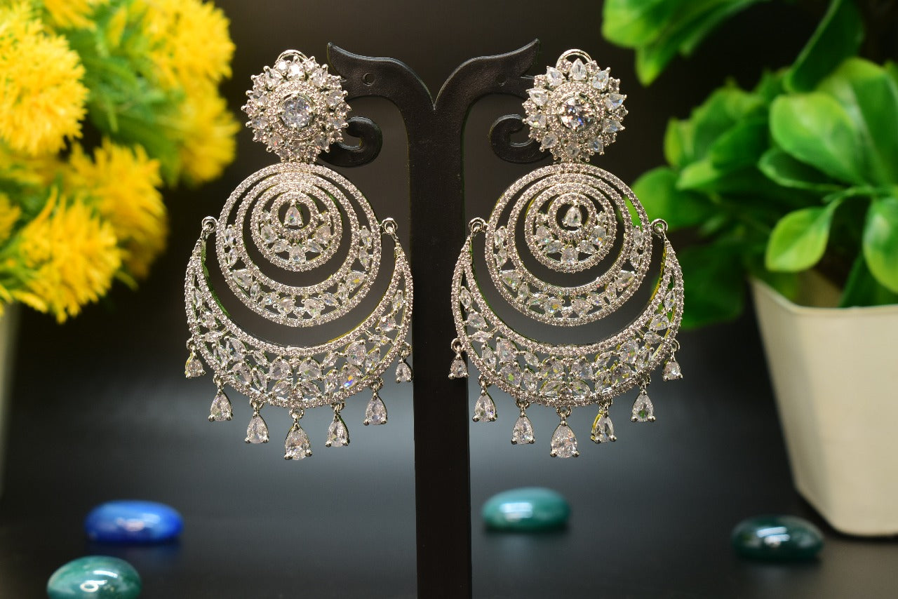 AD Pair of silver Chandelier earrings on a black stand with yellow flowers and green plants in the background