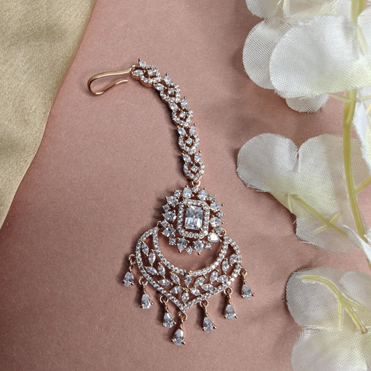 Rose gold Mangtikka and AD diamond jewelry piece on a pink surface with white flowers