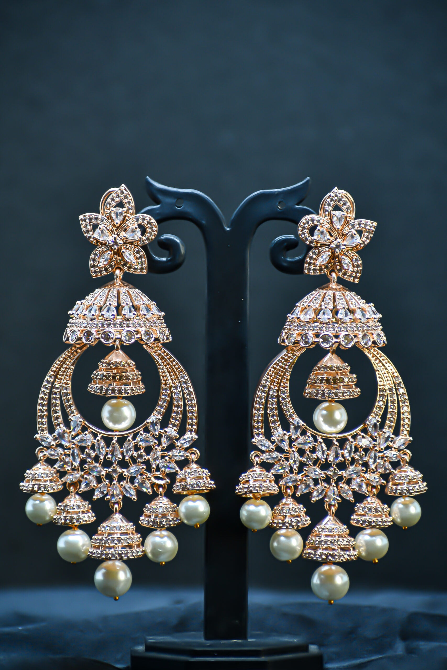 A pair of rose gold-plated zirconia jhumka earrings displayed on a stand against a black background.