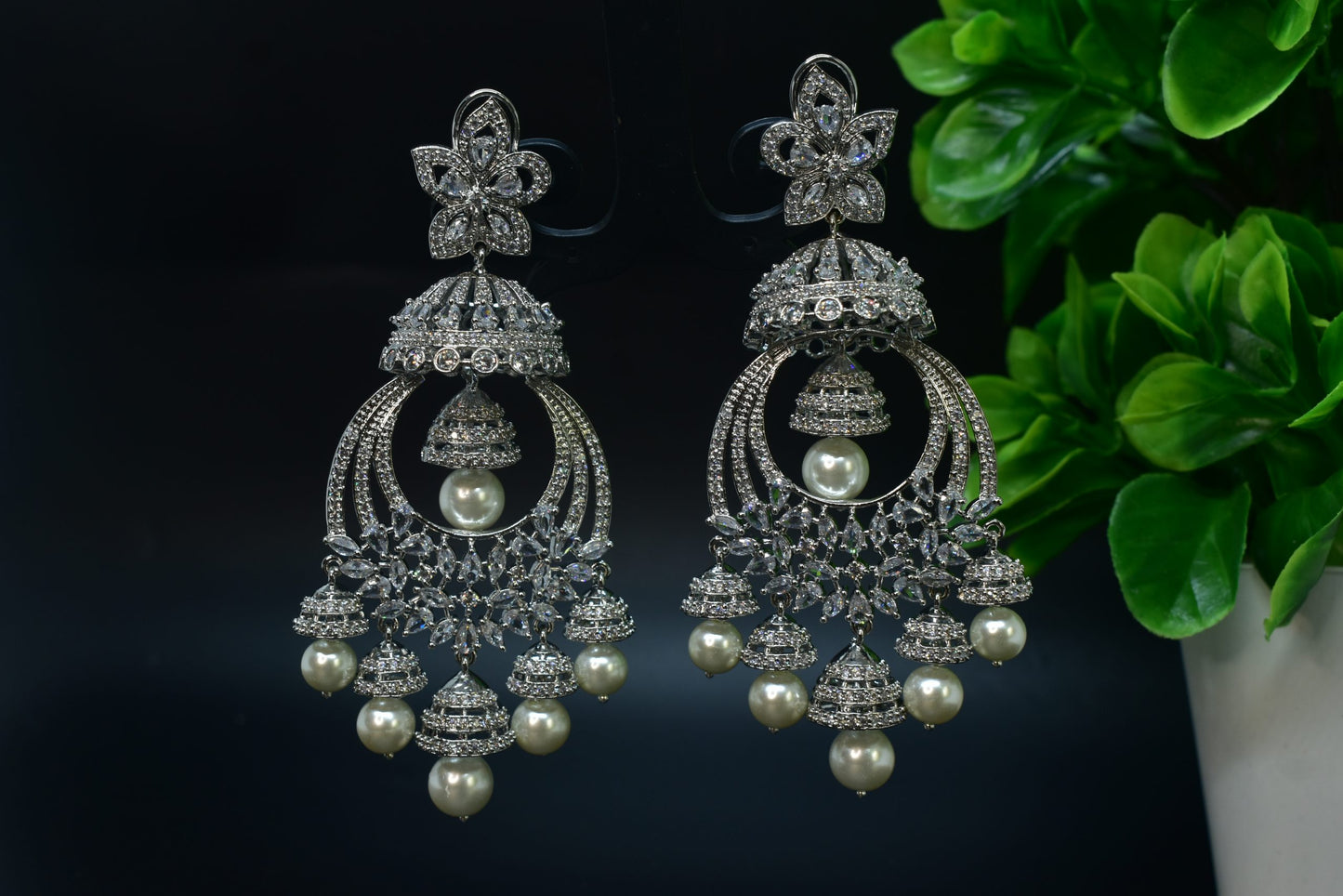 A pair of silver-plated zirconia jhumka earrings displayed on a stand against a black background.